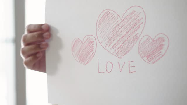 Woman showing a white paper with coloring red heart shape and a word love by crayon