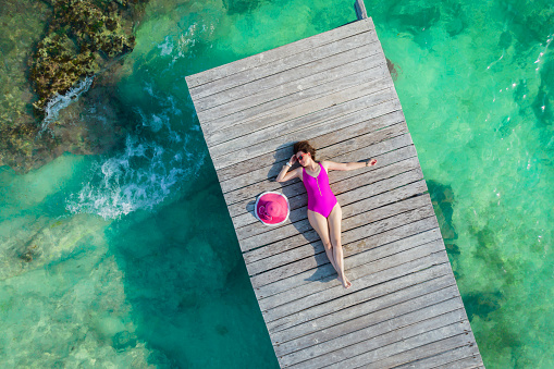 Aerial view of woman laying on wooden pier at sunny summer day in Cancun, Mexico, top view. Young sexy woman wearing bright swimsuit in summertime in Caribbean