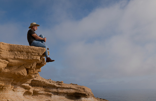 Adult man in cowboy hat sitting on cliff against sky. Almeria, Spain