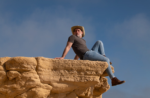 Adult man in cowboy hat sitting on cliff against sky. Almeria, Spain