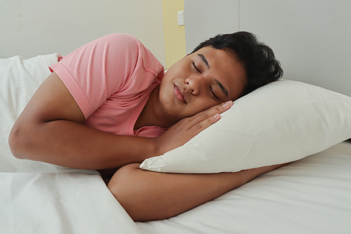 Portrait of Asian man in pink t-shirt smiling while sleeping with side position in the bedroom