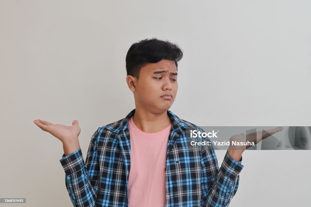 confused Asian man choosing between two different options Portrait of confused Asian man in casual shirt choosing between two different options, holding two hands with empty space. Decision and choice concept Teenager Stock Photo