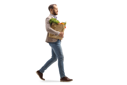 Full length profile shot of a young man carrying a grocery bag and walking isolated on white background