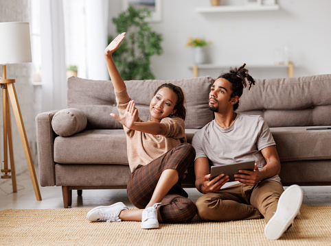 Young lovely african american family couple sitting on floor with digital tablet and discussing new apartment design project or interior design ideas, ethnic man and woman planing home renovation