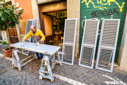 Rome, Italy, January 19 --A craftsman repairs and paints some shutters outside his shop along an alley in the Rione Monti (Monti District), in the historic heart of Rome. The Monti district is a popular and multi-ethnic quarter much loved by the younger generations and tourists for the presence of trendy pubs, fashion shops and restaurants, where you can find the true soul of the Eternal City. This district, located between the Esquiline Hill and the Roman Forum, is also rich in numerous Baroque-style churches and archaeological remains from the Roman era. In 1980 the historic center of Rome was declared a World Heritage Site by Unesco. Wide angle image in high definition format.