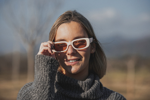 A closeup of a Mexican model wearing sunglasses looking up to the sun. She is also wearing earrings and a black top.