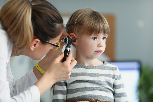 Female pediatrician looking at ear of little girl using otoscope in clinic. Hearing loss diagnosis in children concept