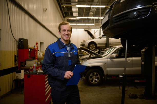 istock Professional portrait of a young Caucasian auto engineer technician mechanic in uniform smiling with a toothy smile looking at camera while standing in repair shop with clipboard in his hands 1368173214