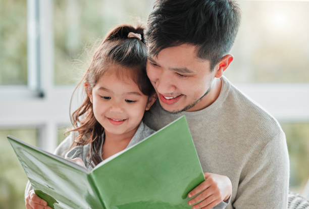 снимок маленькой девочки, сидящей на коленях у отца с книгой - family reading book child стоковые фото и изображения