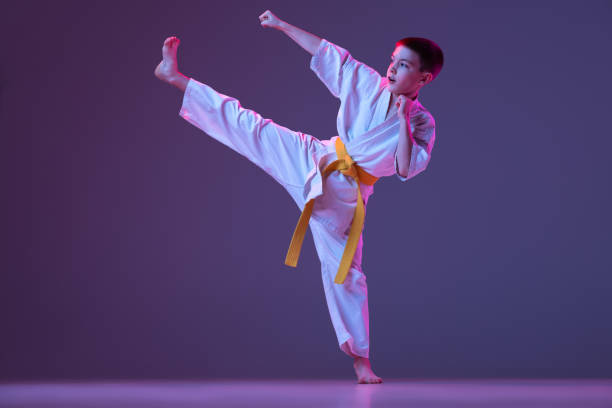 istock Dynamic portrait of kid, young male taekwondo, karate athletes in doboks doing basic movements isolated on purple background in neon. Concept of sport, martial arts 1368177346