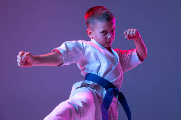 istock Dynamic portrait of kid, young male taekwondo, karate athletes in doboks doing basic movements isolated on purple background in neon. Concept of sport, martial arts 1368177383