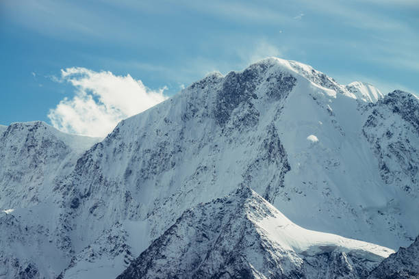 istock Great view to high snowy mountain peaked top with low cloud under cirrus clouds in sky. Low clouds on big snow covered mountains with sharp pinnacle in sunshine. White-snow pointy peak in sunlight. 1368180342