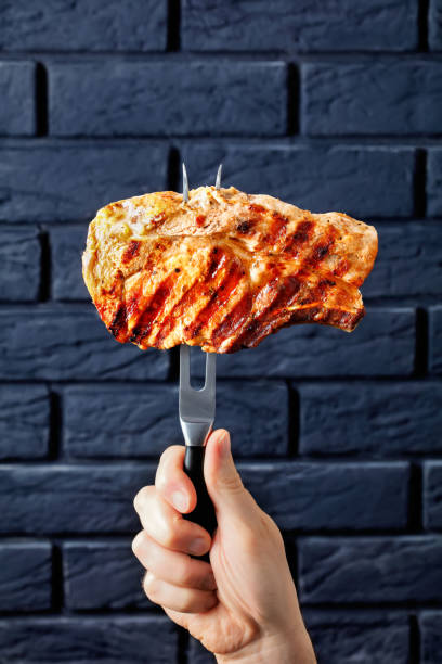 carving fork with speared grilled pork chop hand holds carving fork with speared grilled pork chop on it with a black brick wall at the background carving set stock pictures, royalty-free photos & images
