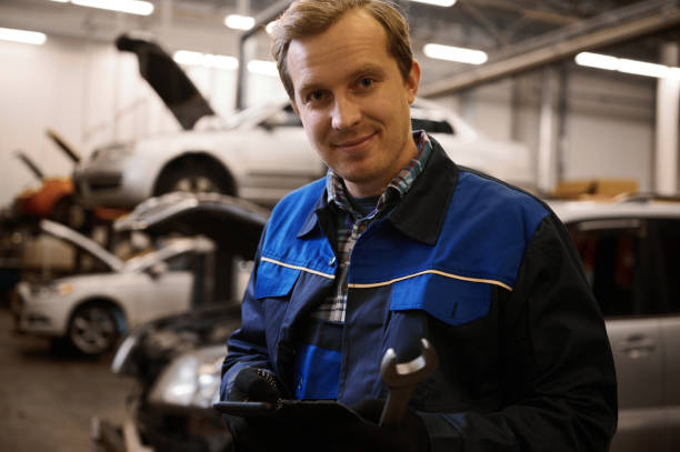 istock Handsome smiling Caucasian auto mechanic, technician in uniform, holding a wrench and standing against lifted cars with open hood in the repair shop 1368186747
