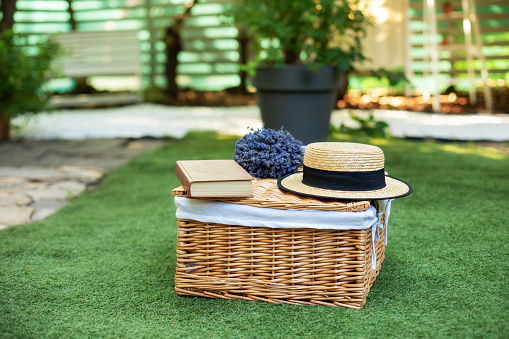 Wicker picnic basket  with flowers on grass in garden. Weekend concept. Picnic basket with a book and a bouquet of lavender on the grass. Cozy Picnic lunch outdoors on the lawn on a sunny day in park.
