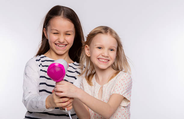 deux belles filles de 6 à 8 ans jouent avec le sèche-cheveux. les filles s’amusent à passer du temps ensemble et à sécher les cheveux sur fond blanc - female 8 9 years child excitement photos et images de collection