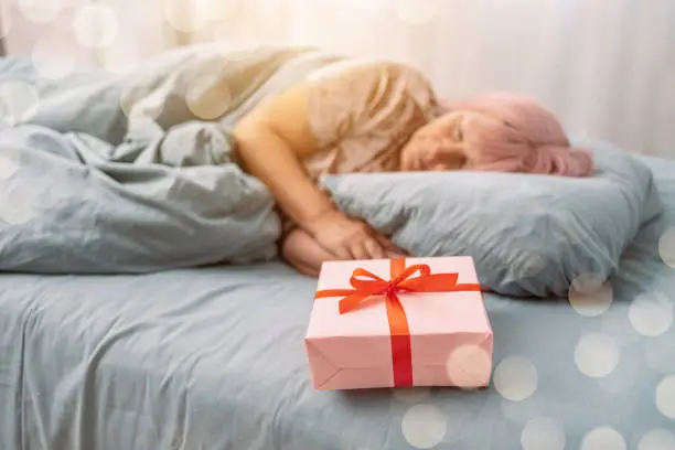 Woman in red sleep mask sleeping in bed near paper gift box with red satin ribbon decor. Christmas, New Year, Valentine's Day and birthday concept