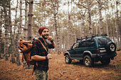 Millennial man hipster with beard wearing backpack, holding axe and firewood in forest with car on background.