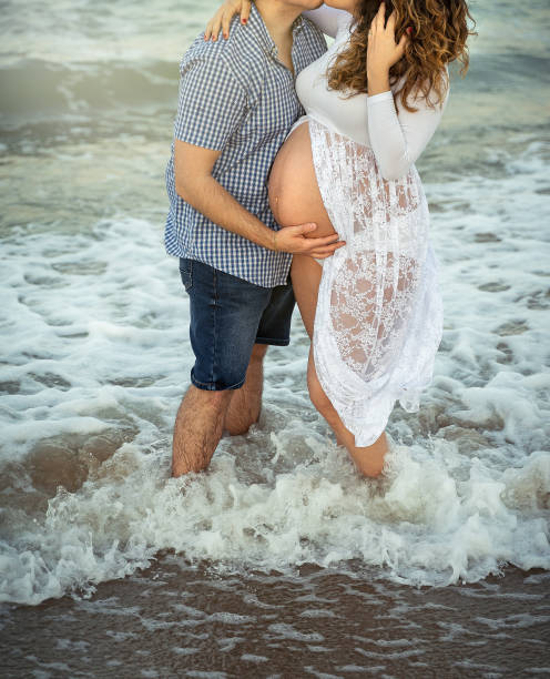 vertical shot of couple kissing with pregnant female in white dress by the beach - human pregnancy couple prenatal care heterosexual couple imagens e fotografias de stock