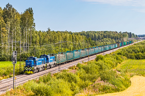Utti, Finland - August 7, 2019:  trainload of harvested cut timber on way to mill passing farmland and plantation trees alongside double-track electrified main railway line. Front view with Fenniarail Dr18 diesel locomotives in foreground with log wagons behind. Train is operated by Fenniarail, private Finnish freight rail operator with timber products from Russia.