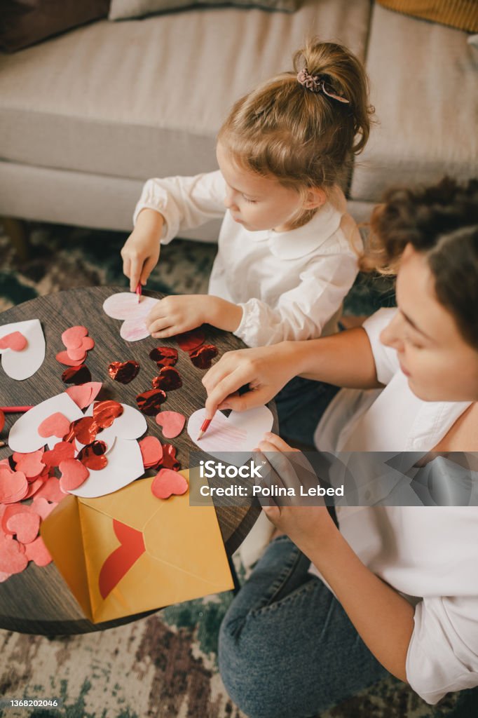 Mother and daughter making Valentine's day cards using color paper, scissors and pencil, sitting by the table in cozy room Mother and little daughter making Valentine's day cards using color paper, scissors and pencil, sitting by the table in cozy room Valentine's Day - Holiday Stock Photo