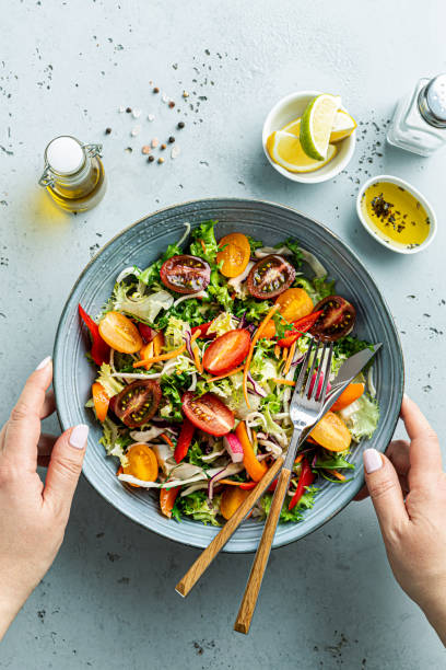 Womanâs hands holding bowl with fresh colorful spring vegetable salad - lunch Fresh colorful spring vegetable salad with cherry tomatoes and sweet peppers. Blue bowl in womanâs hands. Healthy organic vegan lunch captured from above (top view). spring mix lettuce leaves stock pictures, royalty-free photos & images