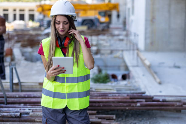 istock Young Female Building Worker is Standing at a Construction Site and Communicate with Coworkers or Friends Using a Smart Phone. 1368215821