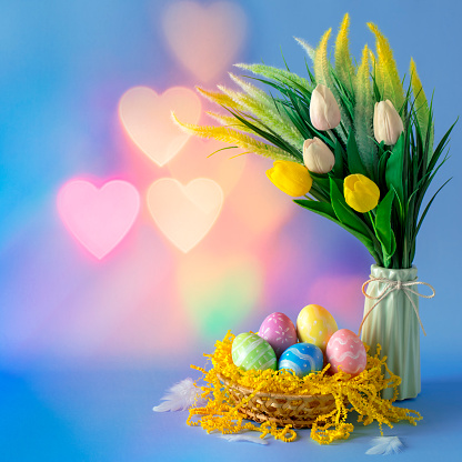 Multi colored hand-painted Easter eggs in a basket with the bouquet of tulips in a vase. Blue background with heart shaped bokeh. It's a REAL bokeh photo, not an illustration or computer filter.
