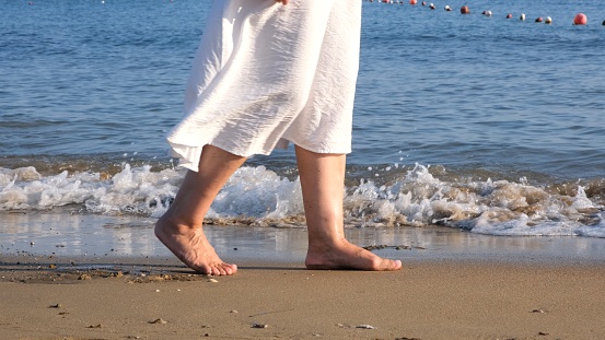 mature female legs are walking along the sandy beach and splashing in the sea on a summer sunny day. Woman walks barefoot on the water close-up.