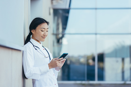 Happy female doctor, smiling and rejoicing, Asian woman uses the phone, during a break near the clinic medical apps, looking at screen, working online, chatting, consulting patient, telemedicine concept
