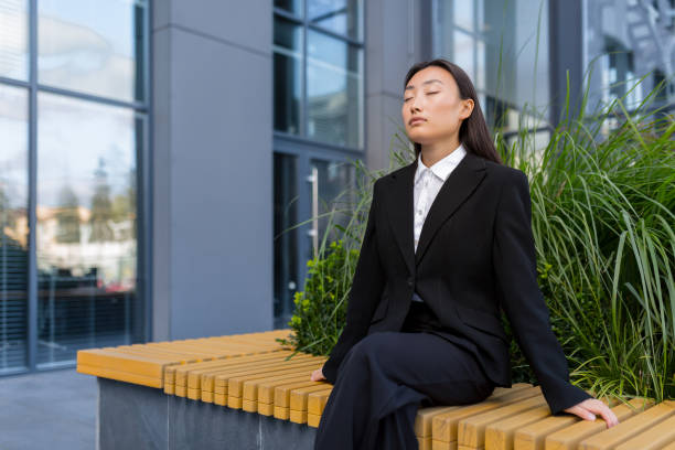 belle femme d’affaires asiatique assise sur un banc se détendant, méditant et effectuant des exercices de respiration - yoga business women indoors photos et images de collection