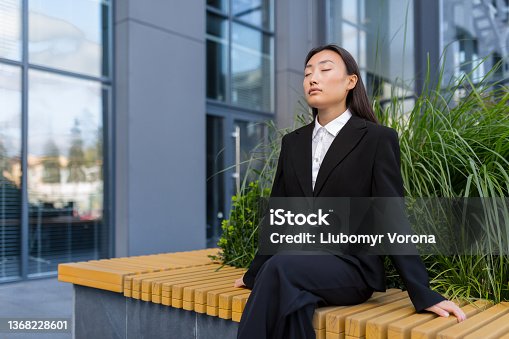 istock Beautiful Asian business woman sitting on a bench relaxing, meditating and performing breathing exercises 1368228601