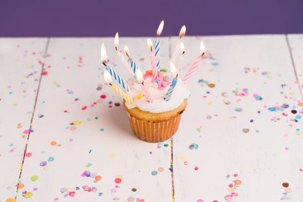 Stock photo shows rich chocolate cupcake topped with rainbow sprinkles and a glowing birthday candle sits atop a muffin, creating a festive dessert scene. The vibrant colors of the sprinkles contrast beautifully with the luxurious velvet red background, adding a touch of elegance and celebration