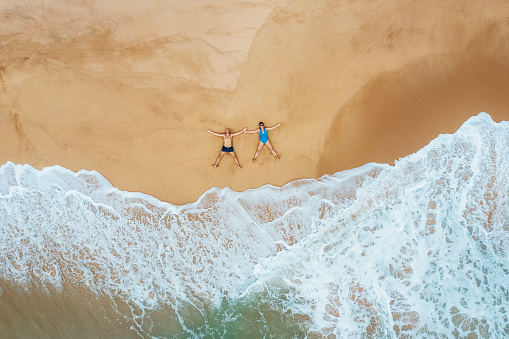 Aerial photo of a smiling couple looking UP dressed swimsuit on the sandy ocean shore and sun tanning. Soft waves on Tangalle beach on Sri Lank island. Exotic carefree vacation top view concept