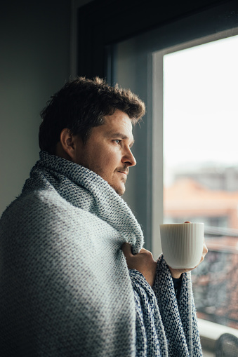 Sick man with blanket looking through the window with a cup of tea