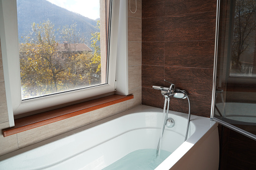 Pure white bathtub with water near window. Bathroom with big window. Mountain view