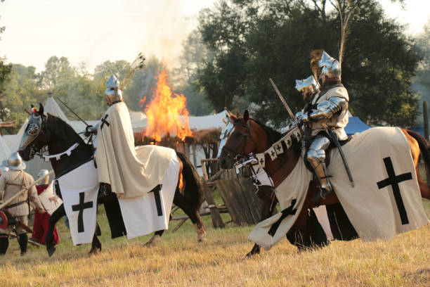 A knight in full armour with helmet, in combat pose with two swords ready to fight. The knight stands by a burning castle, with other knights fighting in the background. The knight is in a fantasy medieval landscape, under a dark and stormy sky.
