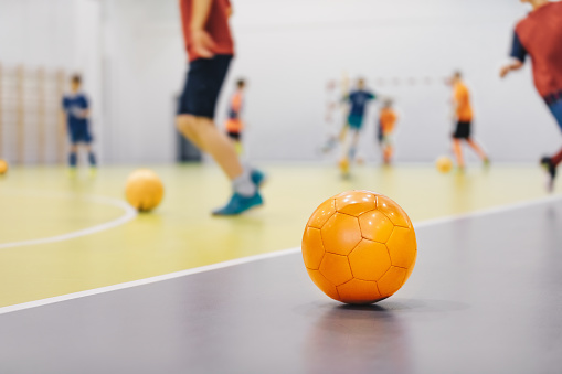 Balón de fútbol sala naranja en el campo de entrenamiento cubierto. Los jugadores jóvenes en deportes practican pelotas de correr en fondo borroso. Clase de deportes escolares para niños photo