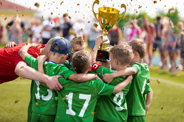 szczęśliwe dzieci wygrywające turniej sportowy. uczniowie stojący z trenerem w kręgu i trzymający złote trofeum. święto zwycięstwa młodzieżowej drużyny sportowej - medal 20s achievement athlete zdjęcia i obrazy z banku zdjęć