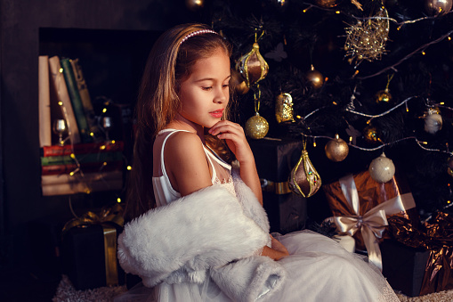 A little girl with brown hair on a dark background in cute dress. Gold and black. Christmas Tree and New Year Decorations