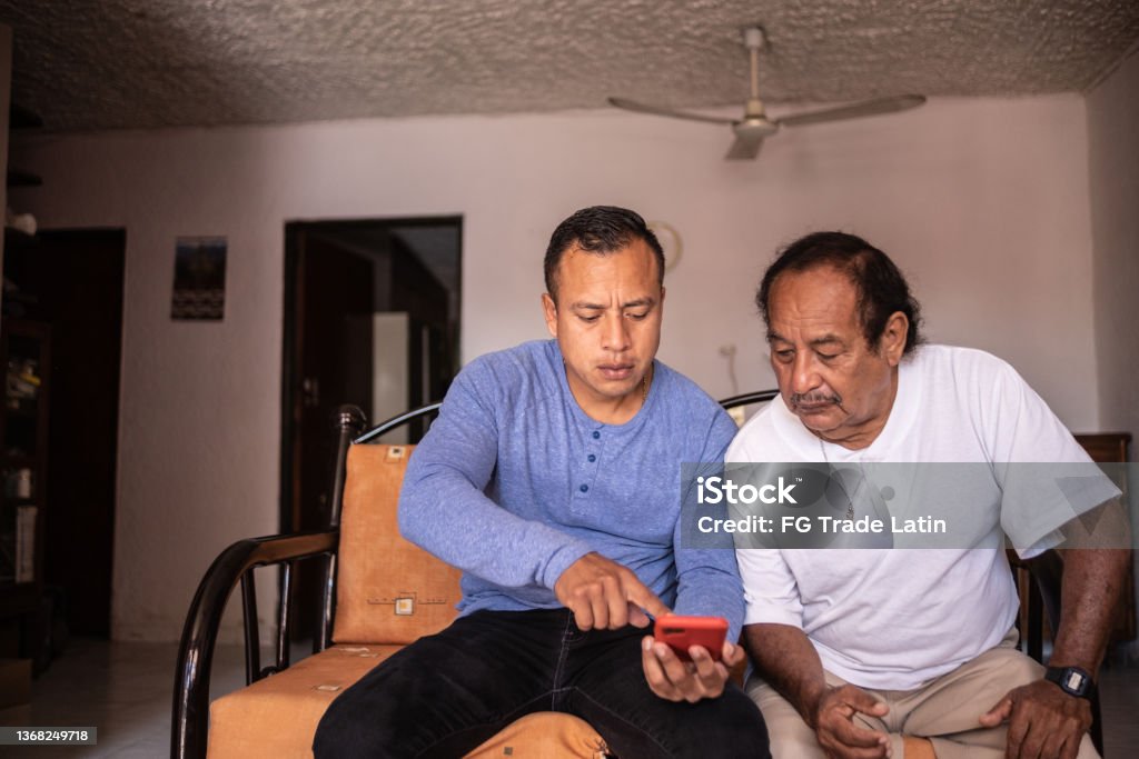 Son helping his father using a mobile phone at home Explaining Stock Photo