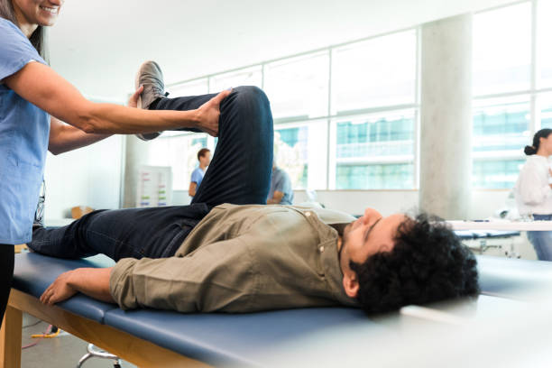Mid adult therapist teaches young man leg stretches The unrecognizable mid adult female therapist stands by the examination table and teaches the young adult man lying down some leg stretches. occupational therapy photos stock pictures, royalty-free photos & images
