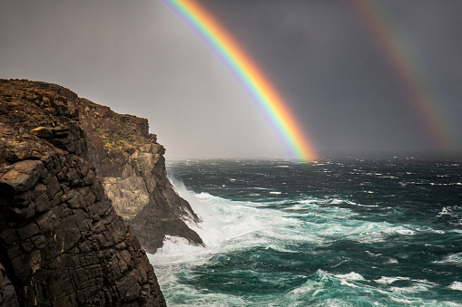 Breathtaking scene of double rainbow behind ocean's horizon near rocky coastline