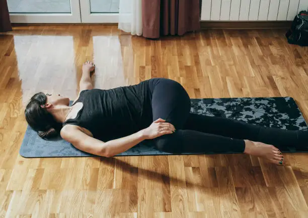 Practicing Reclined Spinal Twist Pose: Beautiful woman in sportswear doing spine rotation exercise on a gray exercise mat in the living room at home.