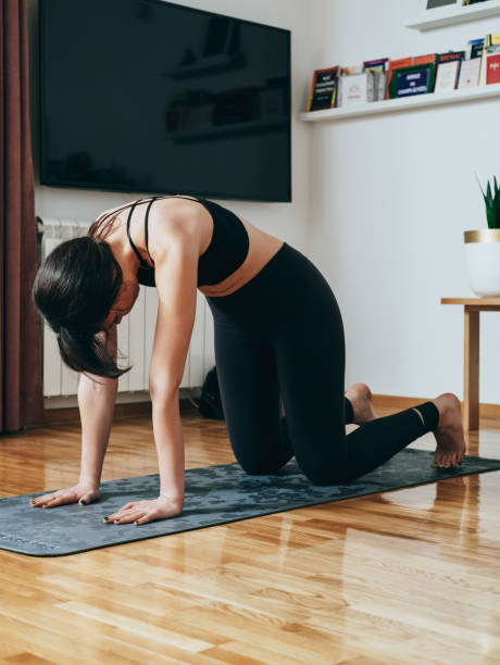 An Anonymous Woman Practicing Yoga in the Morning, Doing Cat Pose Unrecognizable woman in sportswear standing in Marjaryasana pose on a gray exercise mat in the living room at home. cat pose yoga stock pictures, royalty-free photos & images