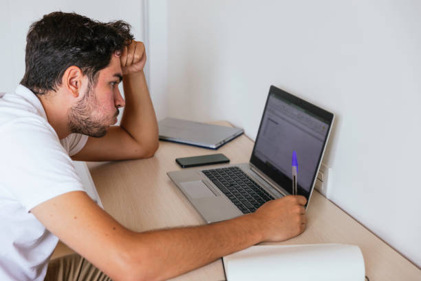 istock Side view of a young man sitting at a desk with a computer taking notes. Concept: teleworking or having online classes 1368276947
