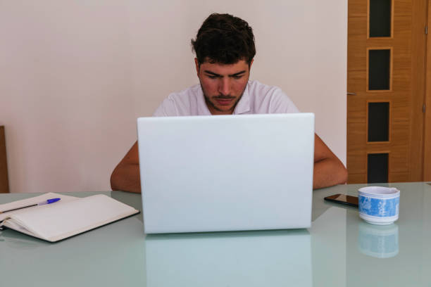 Front view of a concentrated young man working on his computer in his living room, with a coffee, a notebook and his smartphone. Concept: teleworking or having online classes.