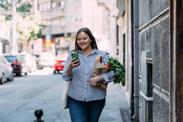 A Smiling Overweight Woman Reading A Message On Her Mobile Phone While Holding A Paper Bag With Groceries In The Other Hand A Caucasian female walking home from shopping while using her smartphone. body confidence stock pictures, royalty-free photos & images