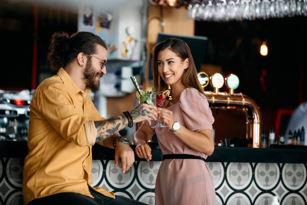 Happy couple toasting with cocktails while celebrating their anniversary at bar counter. Young happy couple celebrating and toasting with cocktails while being on date in a bar. flirting stock pictures, royalty-free photos & images