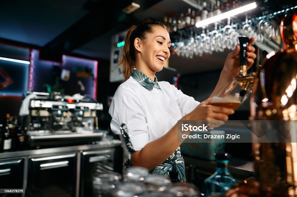 Happy female barista pouring beer from a beer tap while working in pub. Happy female bartender pouring beer draft beer while working at bar counter. Beer - Alcohol Stock Photo